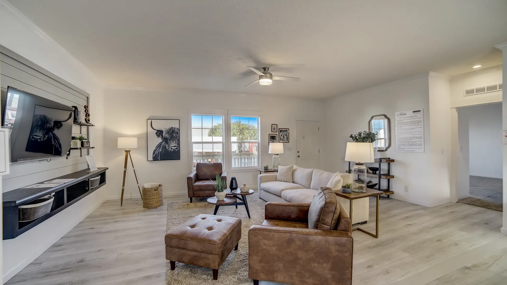 A cozy, modern living room with light wood flooring and neutral tones. Brown leather chairs, a beige sofa, and a TV are arranged around a central coffee table. Wall art depicts animals, enhancing the calm ambiance.