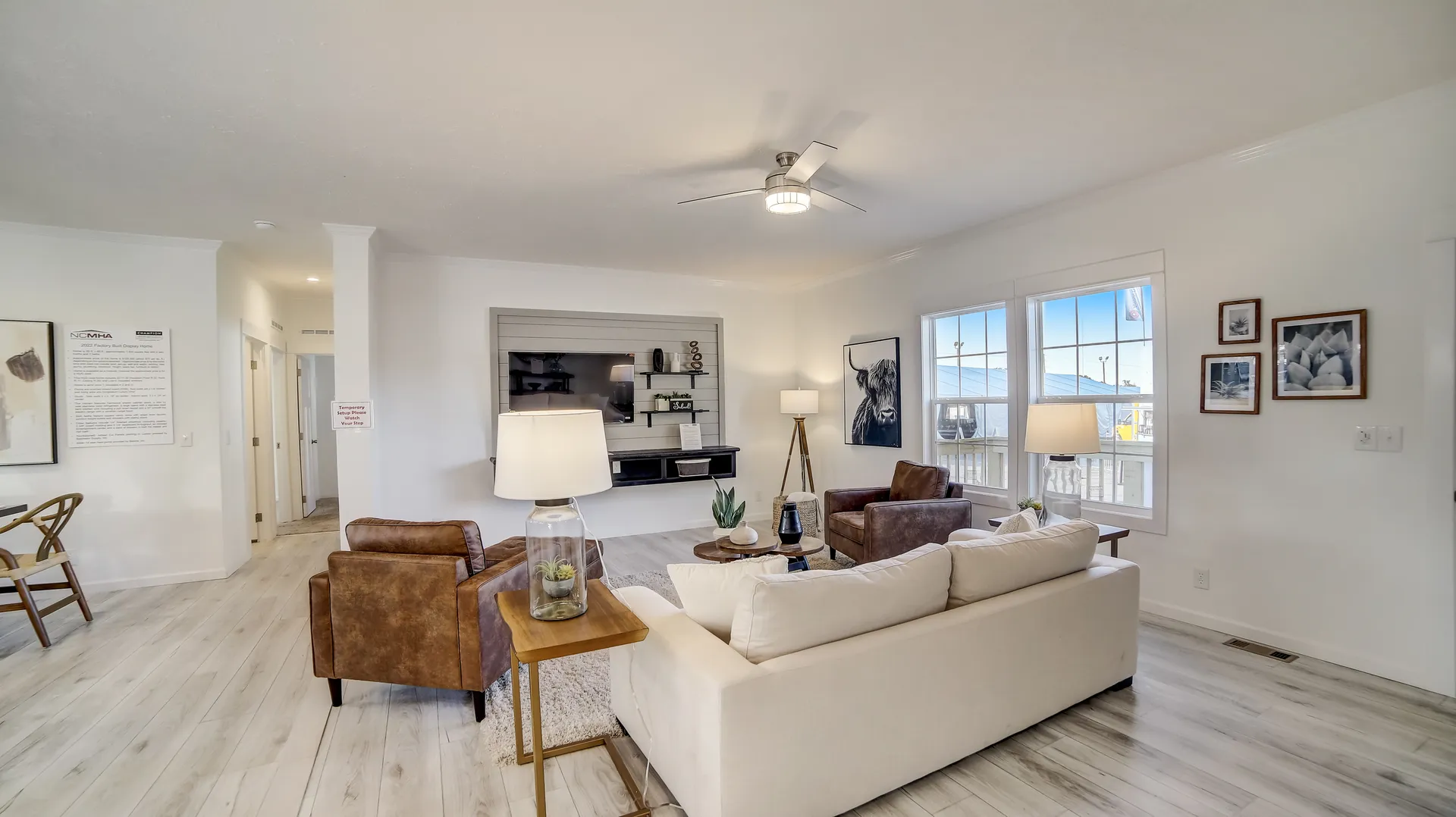 Modern living room with light wood floors, neutral walls, and large windows. Features a cream sofa, brown leather chairs, and a wall-mounted TV. Bright and cozy ambiance.