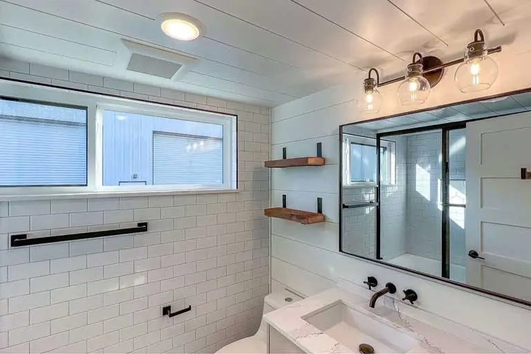 Modern bathroom with white subway tiles and wood accents. Features a large mirror, black fixtures, wooden shelves, and glass light fixtures. Bright, clean, and minimalistic design.