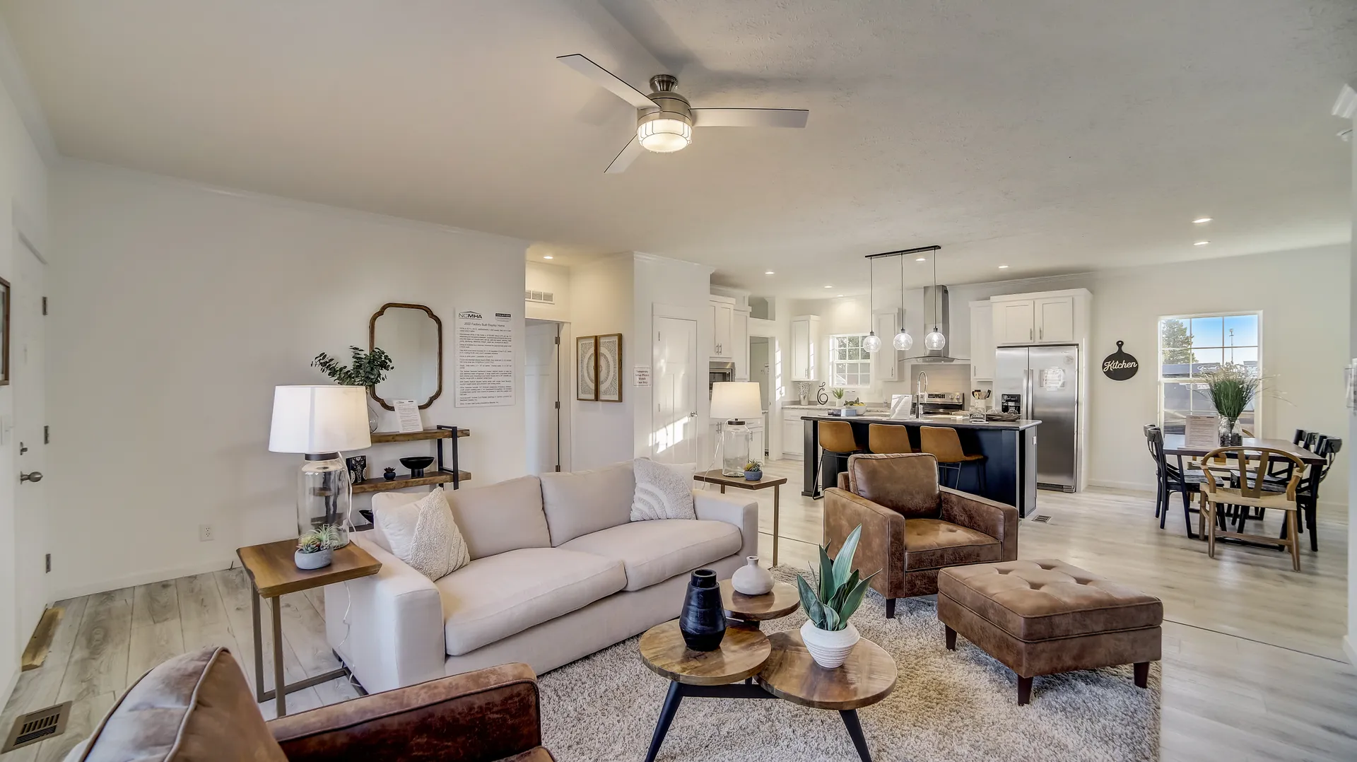 A spacious, modern living area featuring a beige sofa, brown armchairs, and round wooden tables. The open floor plan flows into a kitchen and dining area with sleek, minimalist decor, creating an inviting, cozy atmosphere.