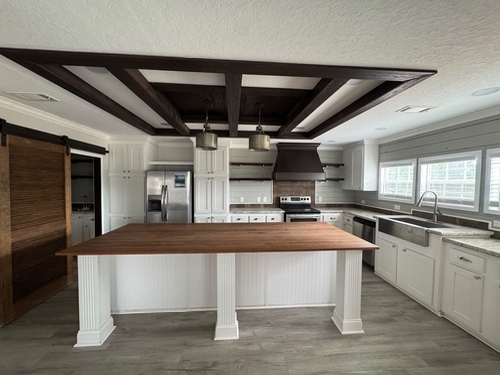 Spacious kitchen with a large wooden island, modern stainless steel appliances, white cabinets, farmhouse sink, and dark ceiling beams. Bright and inviting.