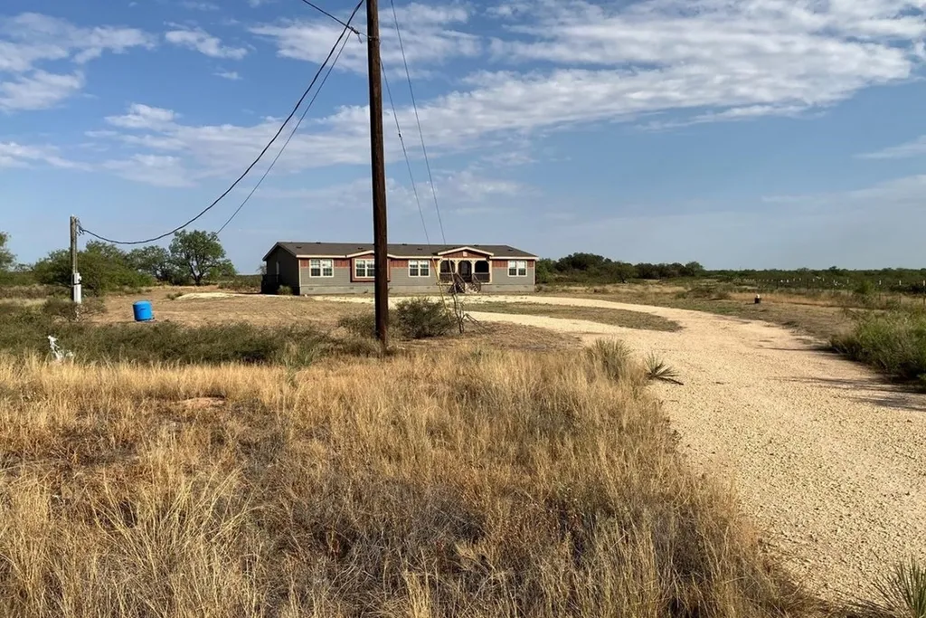 A rural landscape featuring a ranch-style house with a curved gravel driveway, surrounded by dry grass and a wide sky with scattered clouds, evokes a serene and isolated atmosphere.