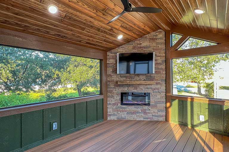Cozy sunroom with wooden ceiling, recessed lighting, and large windows overlooking lush greenery. Features a stone wall with a TV and fireplace.