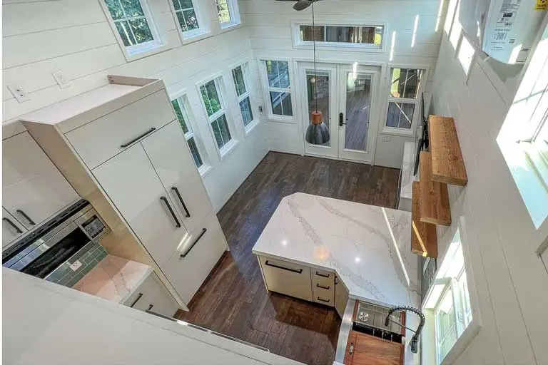Aerial view of a modern kitchen in a tiny house, featuring a marble island, wood flooring, and large windows. Bright, airy space with a cozy vibe.