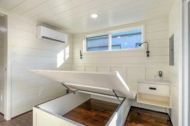A minimalist white room featuring a raised platform bed with storage underneath, a small bedside table, and a window with natural light. Cozy and neat.