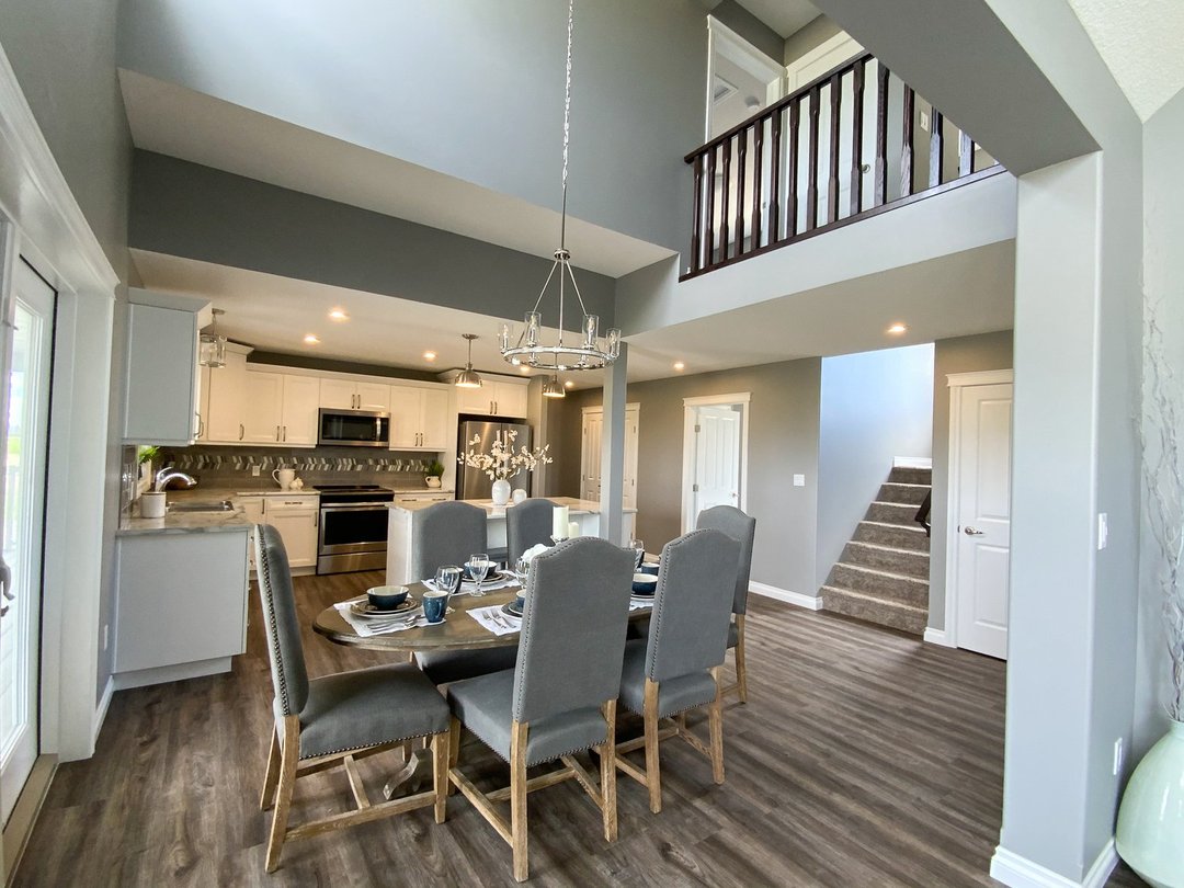Modern dining room and kitchen with high ceiling, gray walls, and hardwood floors. A dining table is set for six, and a chandelier hangs above.