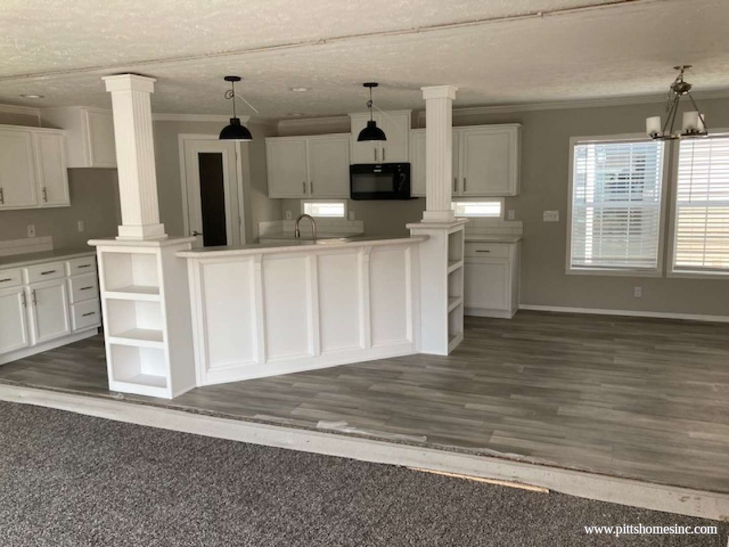 Bright open kitchen with white cabinets, a central island, and hanging black pendant lights. Laminate wood flooring and a cozy, inviting atmosphere.