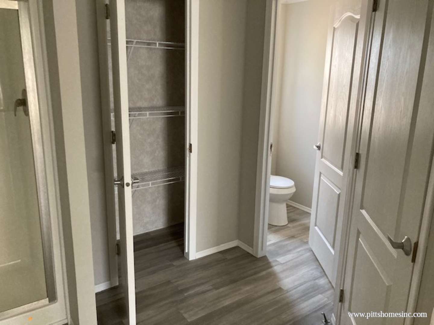 A narrow hallway featuring open closet doors with wire shelving on the left and a closed door on the right. A toilet is visible at the end, suggesting entry to a bathroom. The scene is lit softly, with light gray walls and wood-style flooring, creating a clean, neutral tone.