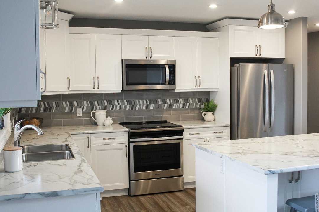 Modern kitchen with white cabinets, marble countertops, and stainless steel appliances, including a fridge and microwave. Gray tile backsplash adds contrast.