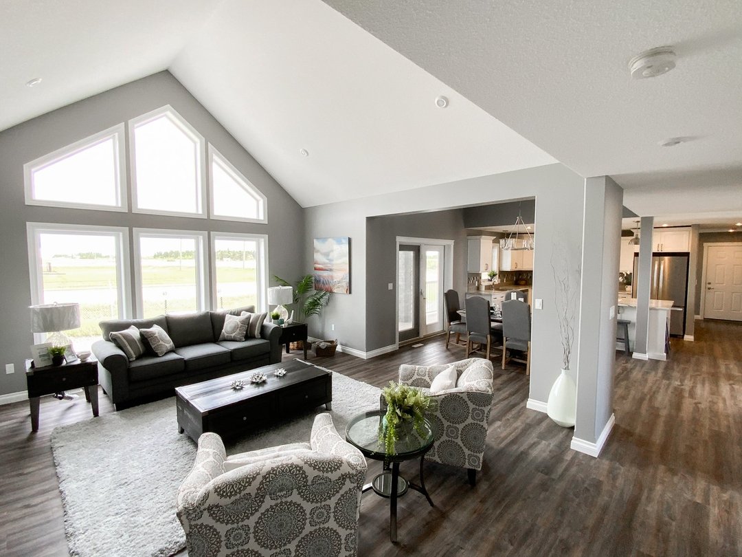 Spacious, modern living room with high ceilings, large triangular windows, and abundant natural light. Features dark furniture, neutral tones, and wooden flooring. Adjacent dining area and kitchen visible in open layout. Cozy and inviting ambiance.