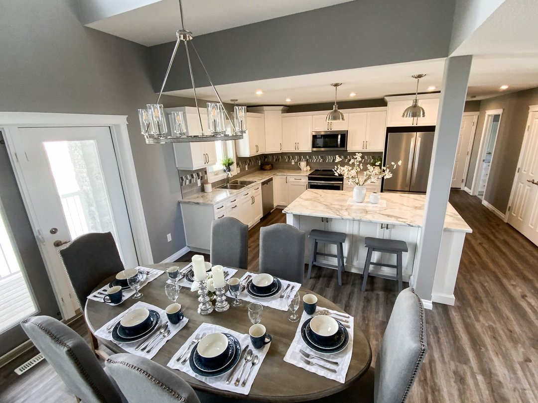 Modern kitchen and dining area with grey walls and hardwood floors. The dining table set for six with elegant dishware. The white kitchen features marble countertops, stainless appliances, and pendant lighting, creating a chic, inviting atmosphere.