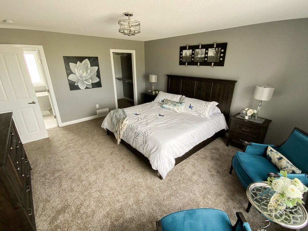 Modern bedroom with a dark wood bed, white bedding featuring bird designs, and teal chairs. Gray walls display flower artwork, exuding a calm ambiance.