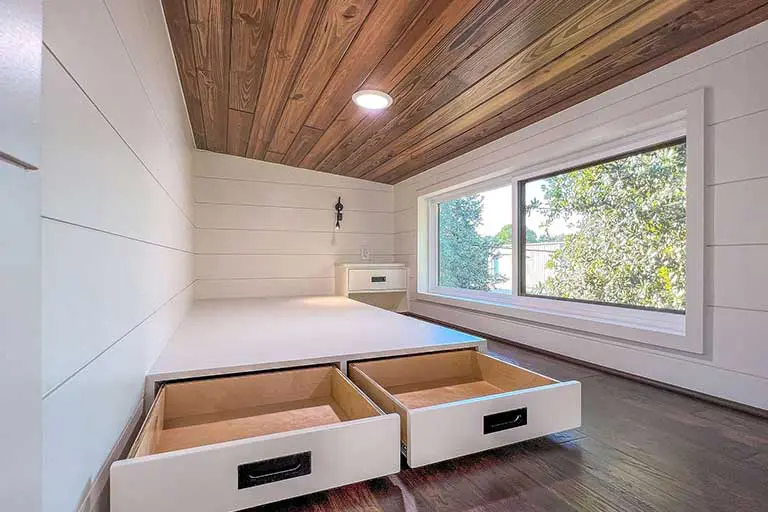 Modern loft space with wooden ceiling, white paneled walls, a large window showcasing greenery, and pull-out storage drawers on a sleek platform. Bright, cozy ambiance.