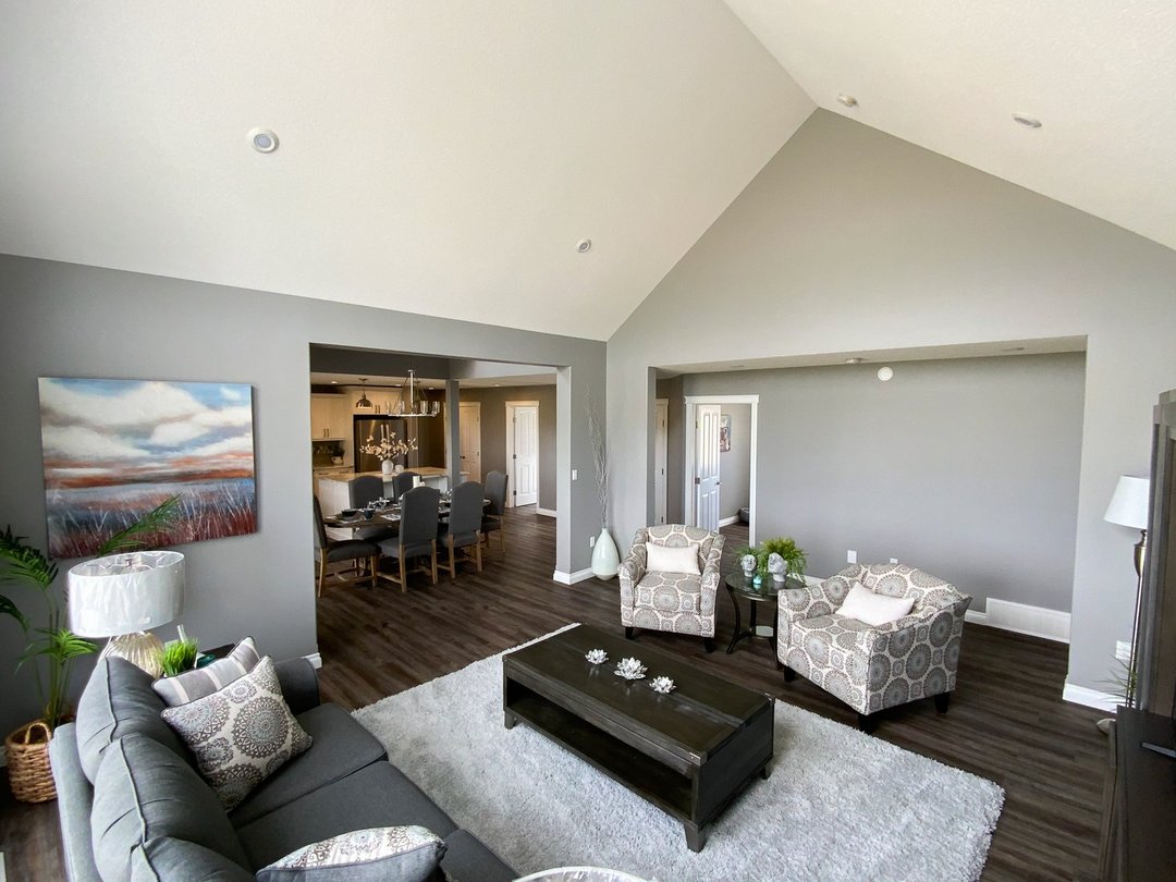 Spacious living room with vaulted ceiling, gray walls, and dark wood floors. Features a gray sofa, patterned armchairs, and a dining area visible. Calm ambiance.