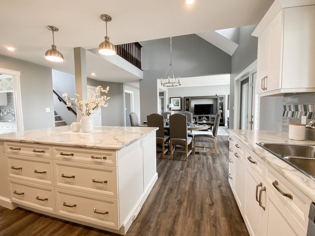 Modern kitchen and dining area with gray walls and wooden floor. White marble island in foreground, elegant dining set and chandelier in background, creating an inviting ambiance.