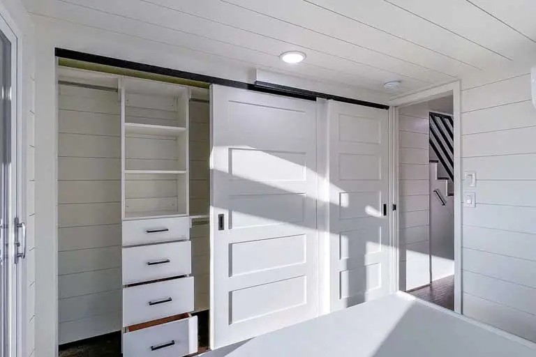 Compact white closet with sliding doors in a modern, bright room. Open shelves and drawers visible. Clean lines create a minimalist, organized feel.