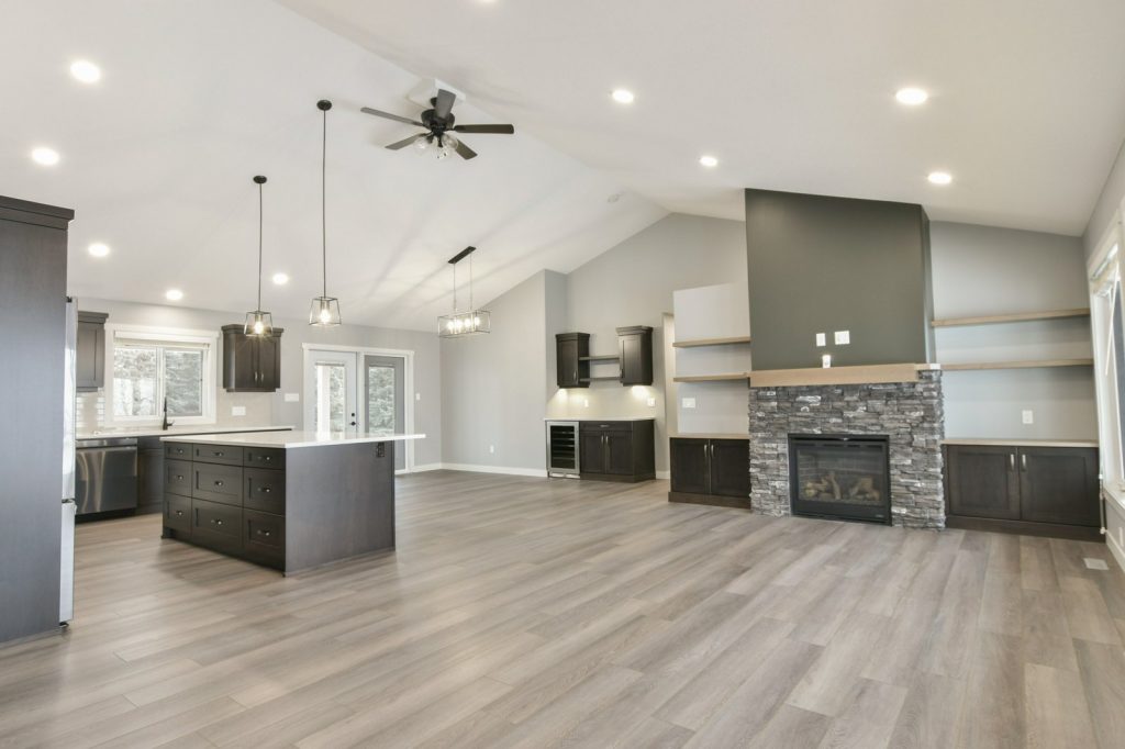 Spacious open-plan kitchen and living area with vaulted ceiling. Features dark wood cabinetry, island, stone fireplace, and modern light fixtures.
