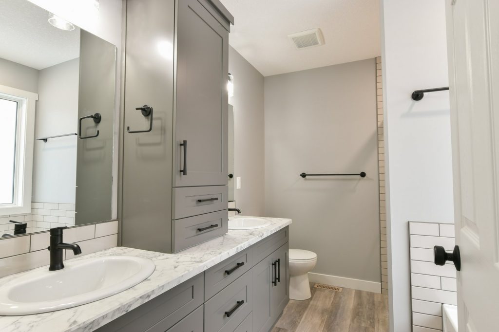 Modern bathroom with gray cabinetry, marble countertop, two sinks, and black fixtures. A toilet and a towel rack are visible, with neutral decor.