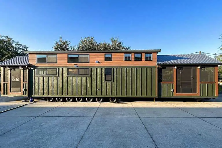 Modern green tiny house on wheels with a wooden accent, featuring multiple small windows and a metal roof, set on a sunny day with trees in the background.