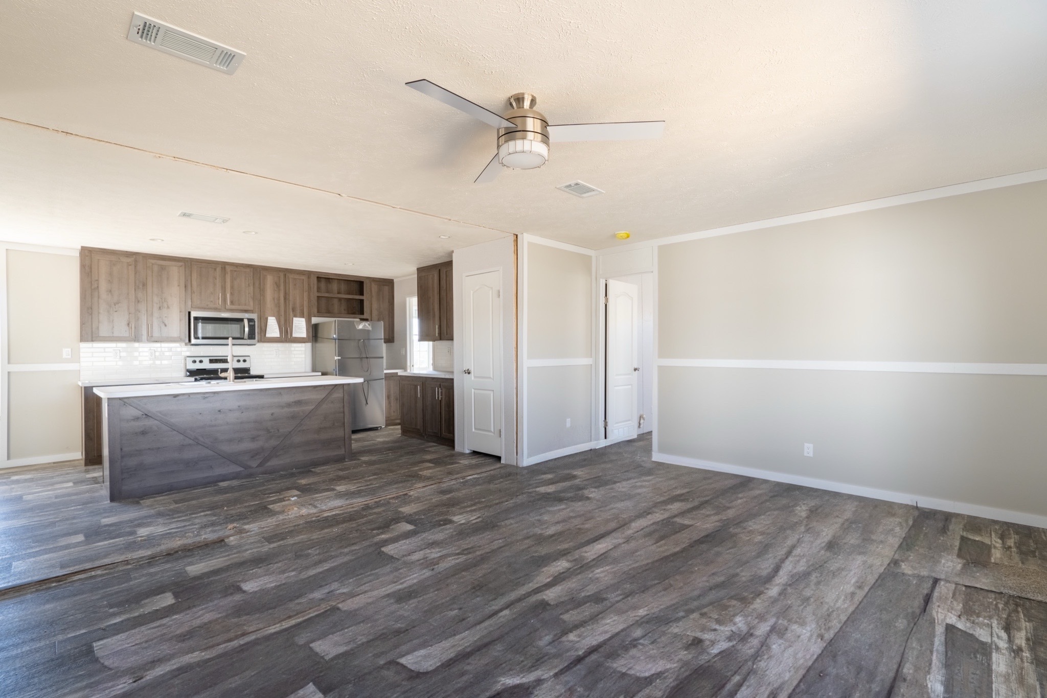 Spacious open-plan room with gray wood flooring, beige walls, and a modern kitchen. Features a ceiling fan and stainless steel appliances, exuding a minimalist style.