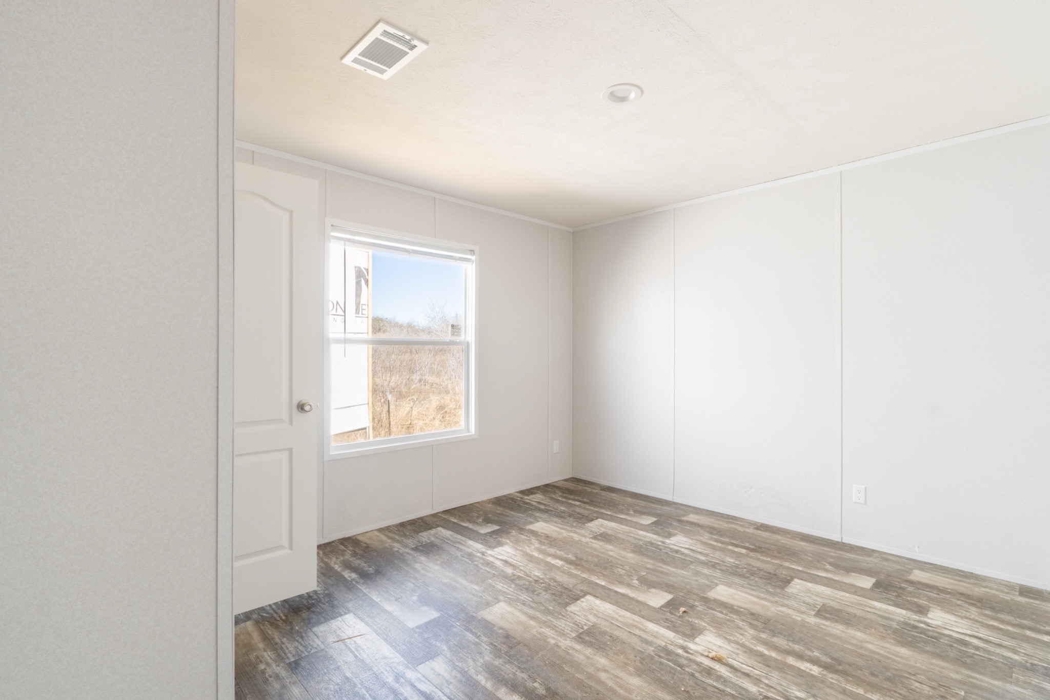Empty room with white walls, a single window allowing natural light, wood-look flooring, and a white door partially open, creating a calm and airy atmosphere.