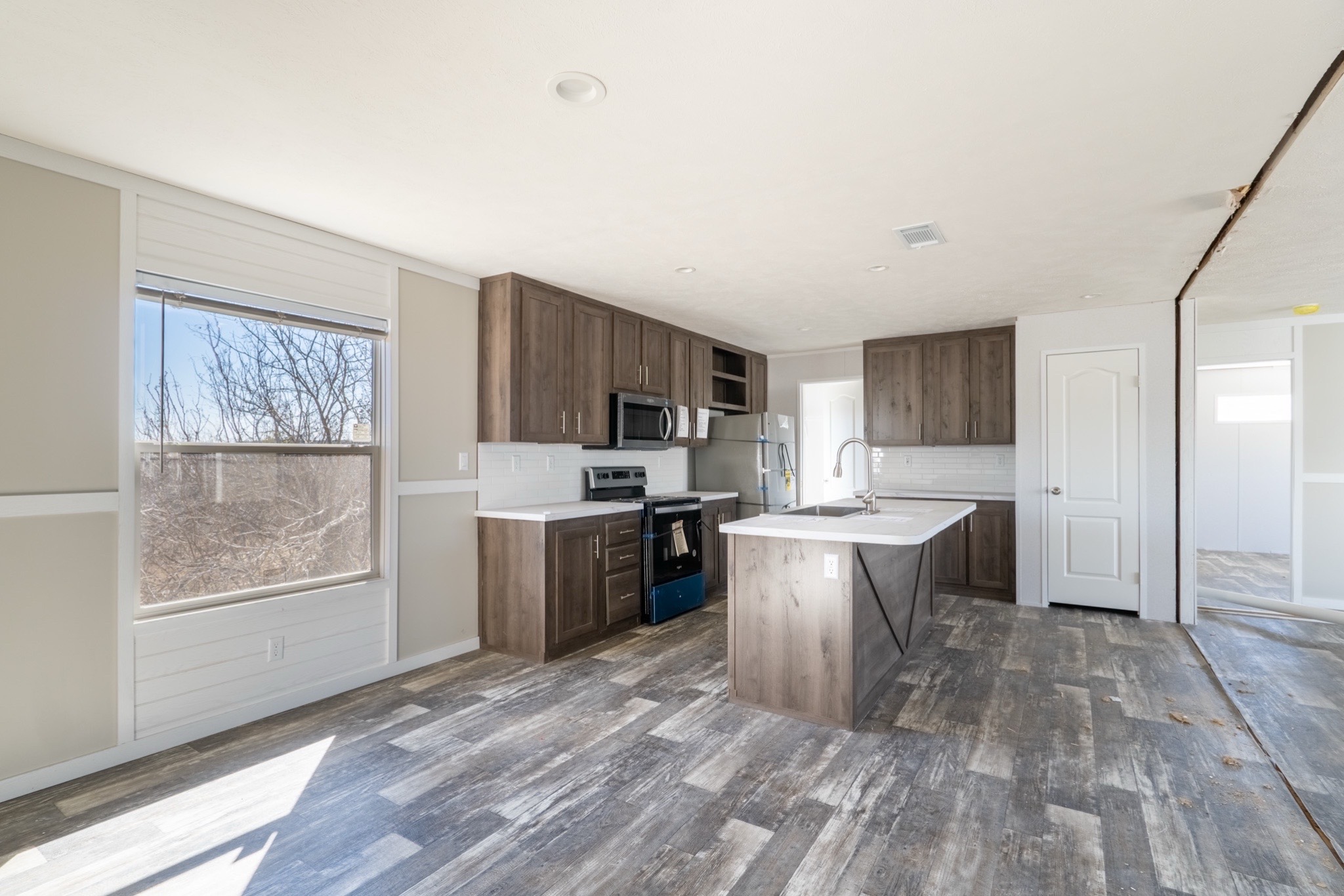 Modern kitchen with rustic wood flooring, sleek dark wood cabinets, and a central island. Sunlight streams through a large window, creating a bright, welcoming atmosphere.