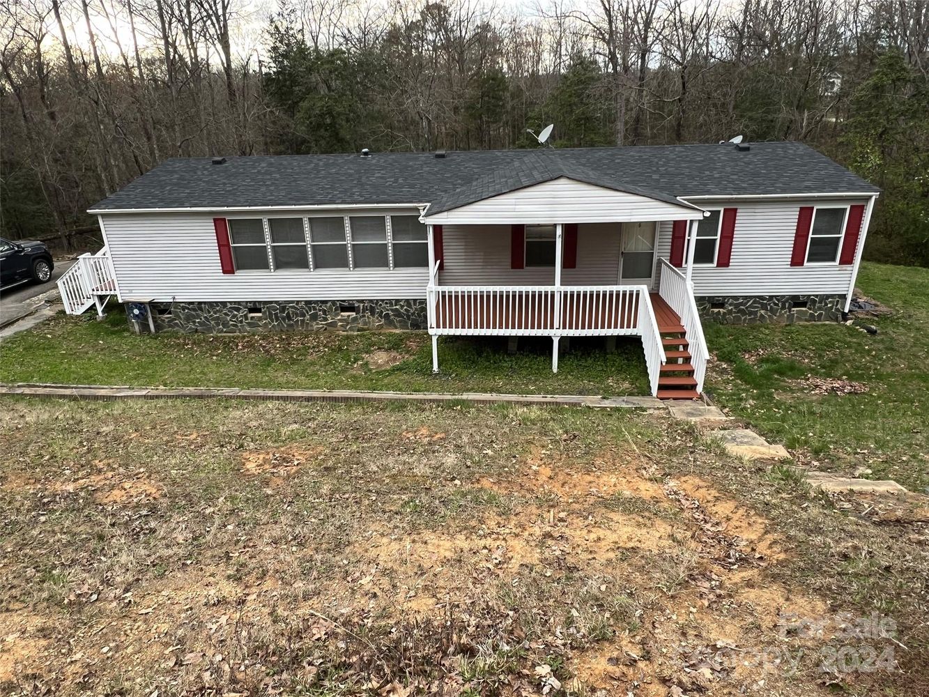 Single-story house with white siding, red shutters, and a black roof, surrounded by trees. It features a front porch with steps and a sloped, grassy yard.