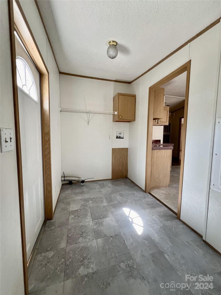 Narrow entryway with gray tile flooring, wood-trimmed walls, a ceiling light, and cabinets. Door is on the left, kitchen visible through an open doorway.