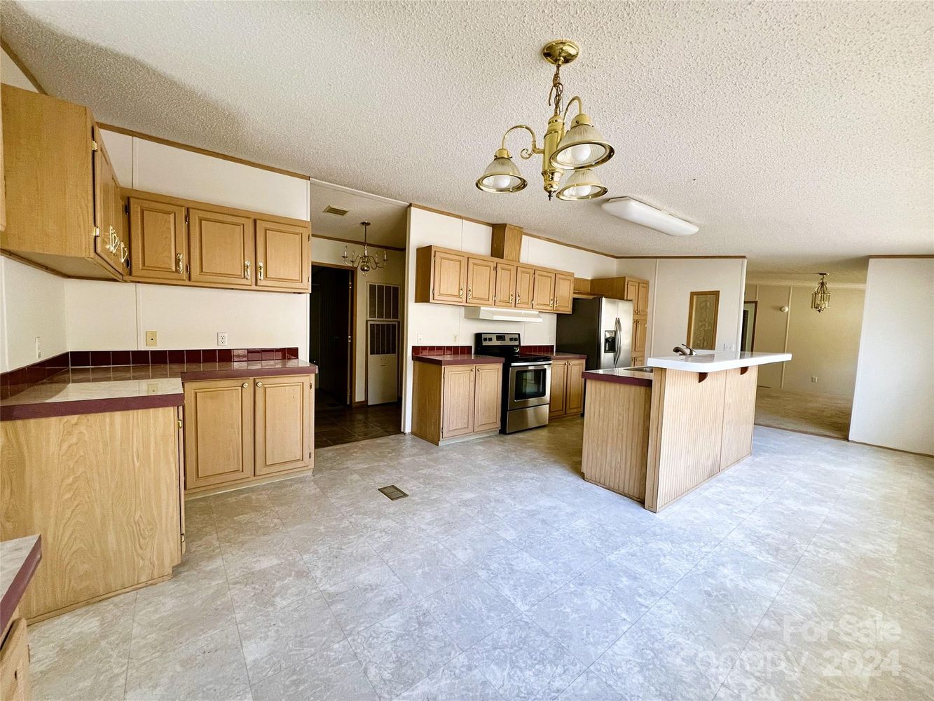 Spacious kitchen with light wood cabinets, red-tiled countertops, and a central island. Stainless steel appliances and beige tile flooring; elegant light fixtures overhead.