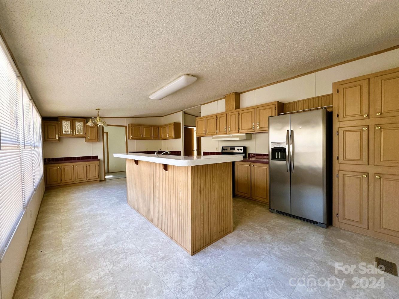 Spacious kitchen featuring wooden cabinets, large island with white countertop, stainless steel fridge, and neutral tile flooring. Bright and inviting atmosphere.