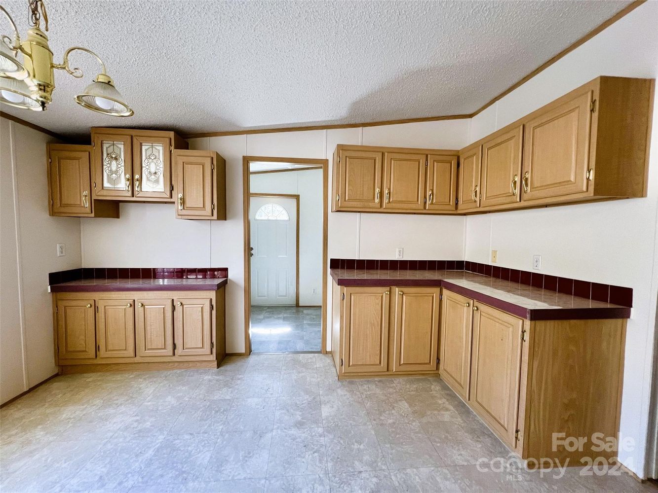 A spacious kitchen with light wood cabinets and brown countertops. It features a simple chandelier, tile flooring, and a door leading to another room.