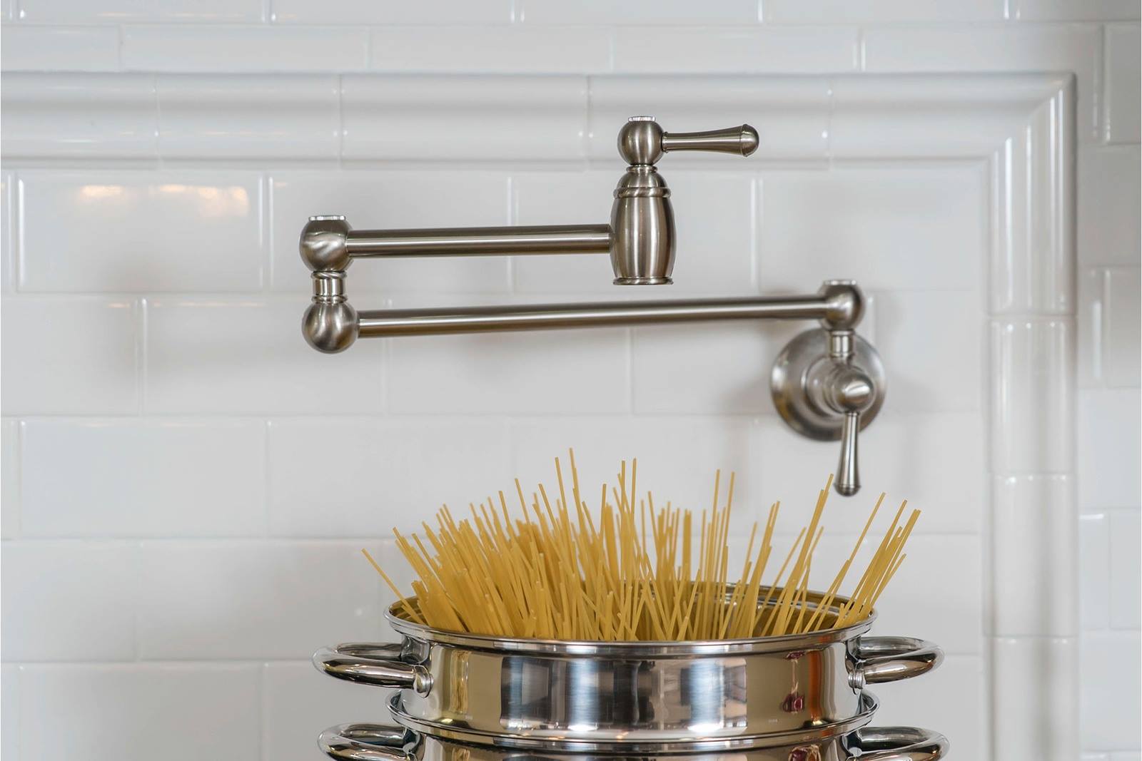 A pot filler faucet overhangs a shiny metal pot filled with uncooked spaghetti. The white tiled backsplash adds a sleek and modern kitchen vibe.