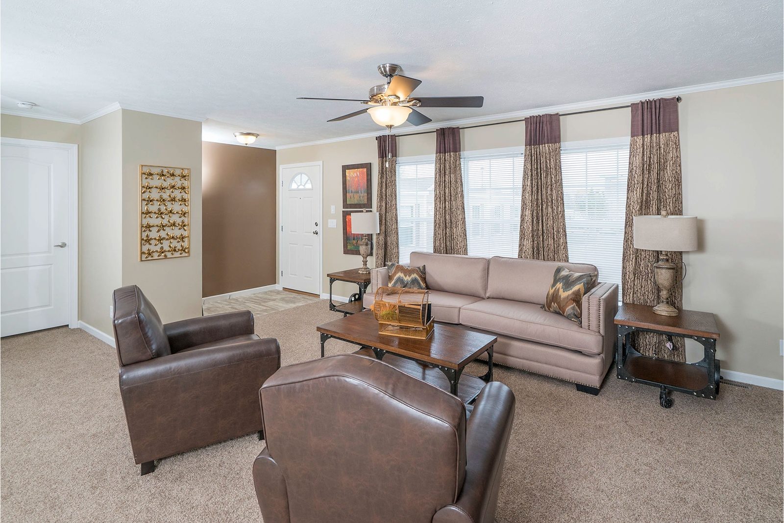 A cozy living room with a beige sofa, two brown leather chairs, and a wooden coffee table. Large window with patterned curtains, soft lighting.