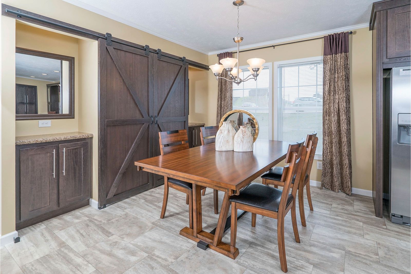 A cozy dining room features a wooden table with six chairs, decorative vases, a chandelier, large windows with curtains, barn doors, and a fridge.