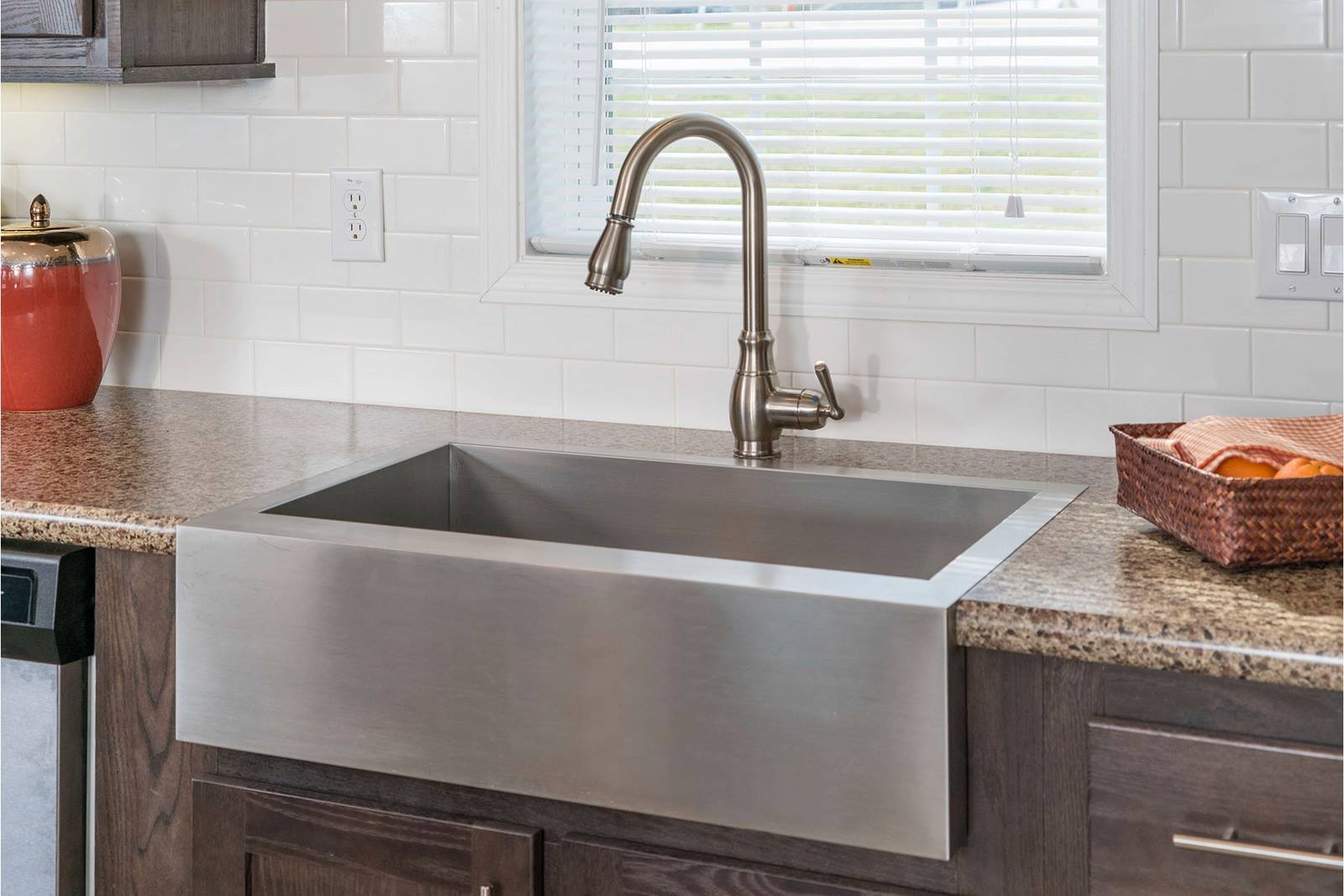 Modern kitchen design with a stainless steel farmhouse sink below a window with blinds. White subway tiles and granite countertops add elegance.