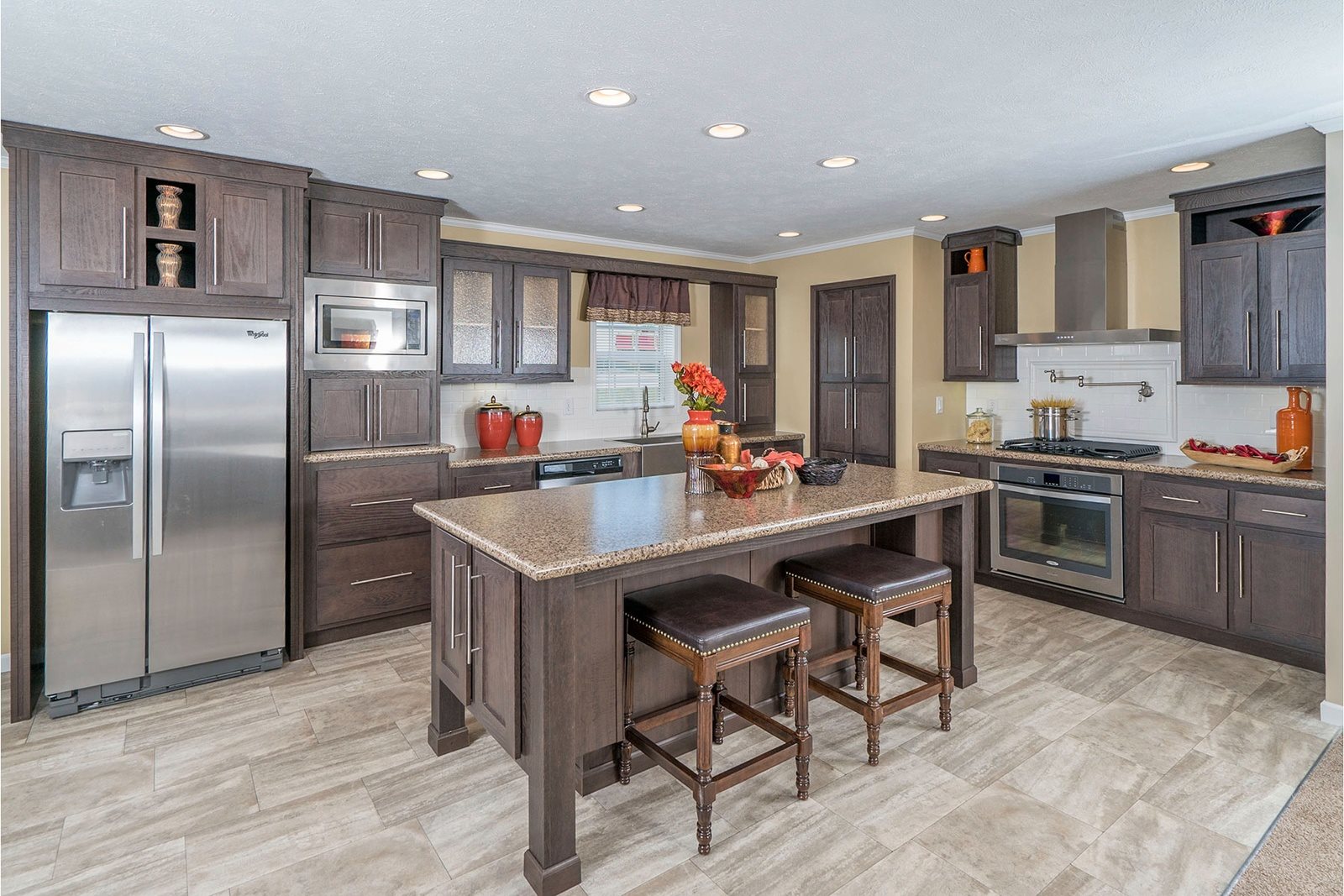 Spacious modern kitchen with dark wood cabinets, stainless steel appliances, and a large island. Two stools sit by the island, decorated with red accents.