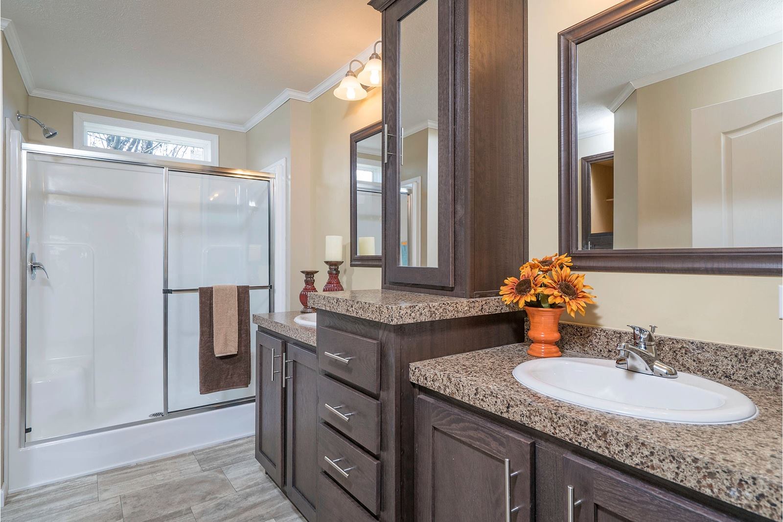 Modern bathroom with dark wood cabinets, granite countertops, and dual sinks. A glass shower enclosure and autumn floral decor create a warm ambiance.