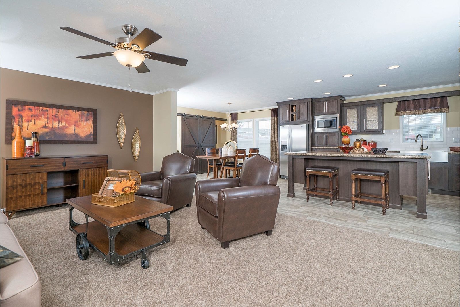 Cozy living area with brown leather chairs, a wooden coffee table, and wall art. Open kitchen has a bar, stools, and dining space, creating a warm, inviting atmosphere.