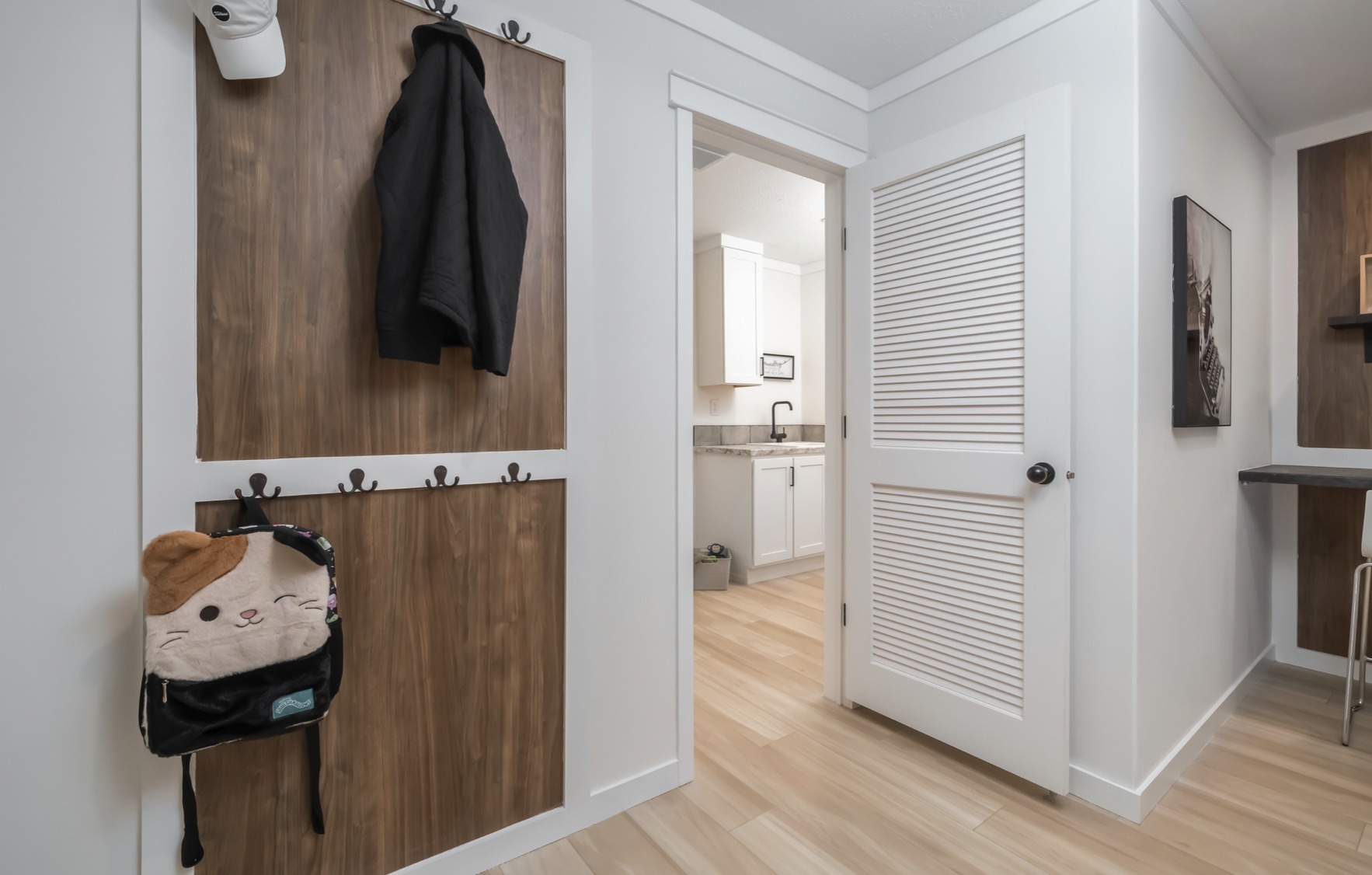 Hallway with light wood floors, wooden panels with hooks on the left holding a black coat and a cute cat-themed backpack. Open door leads to a kitchen.