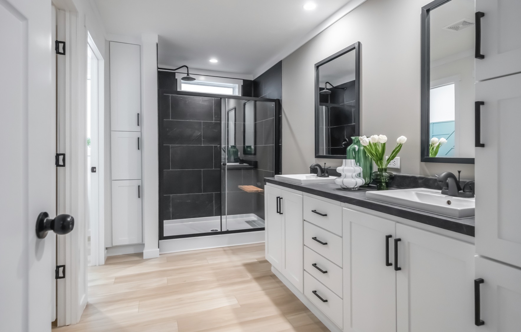 Modern bathroom with dual sinks, black countertops, and white cabinetry. Glass shower with dark tiles on the left. Vase of tulips adds elegance.