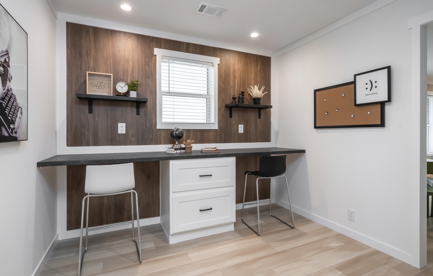 A modern home office with wood accent wall, black shelves, white desk, and barstools. Decor includes plants and books, creating a cozy, minimalist vibe.