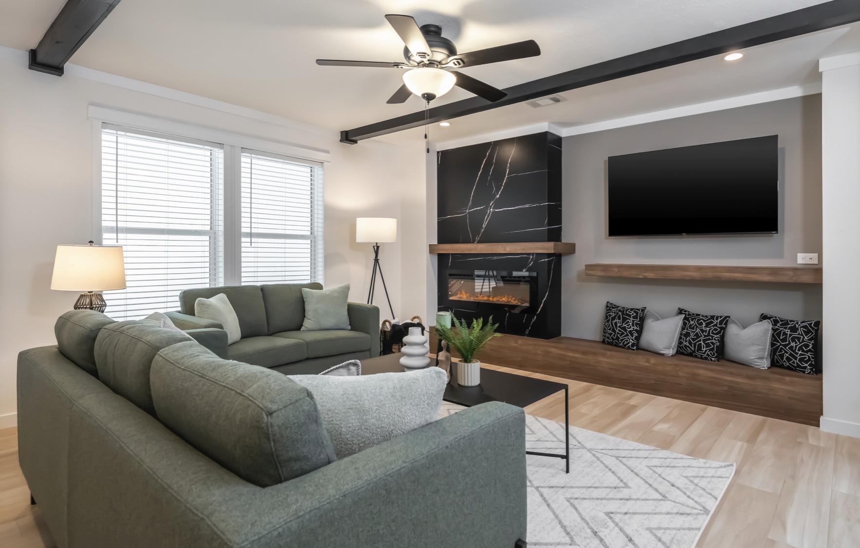 Modern living room with a green sectional sofa, cozy cushions, and a sleek black fireplace. Natural light streams through large windows, creating a warm, inviting atmosphere.