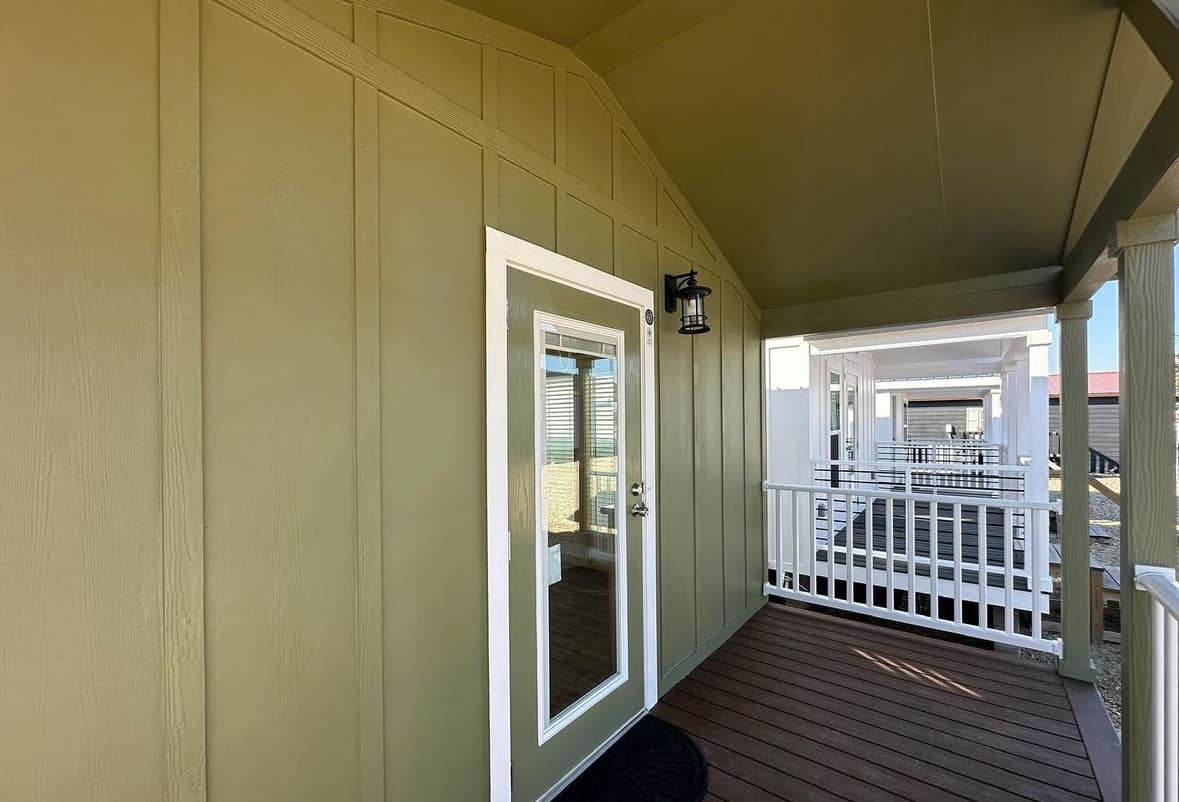 Covered porch with olive-green siding and a glass-paneled door. A lantern-style light fixture is mounted beside the door. White railing lines the porch, leading to other similar structures in the background. The atmosphere feels inviting and modern.