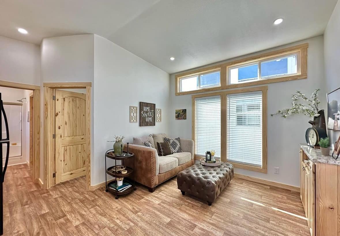 Bright living room with hardwood floors, beige sofa, tufted ottoman, and wooden accents. Sunlight streams through tall windows, creating a cozy atmosphere.