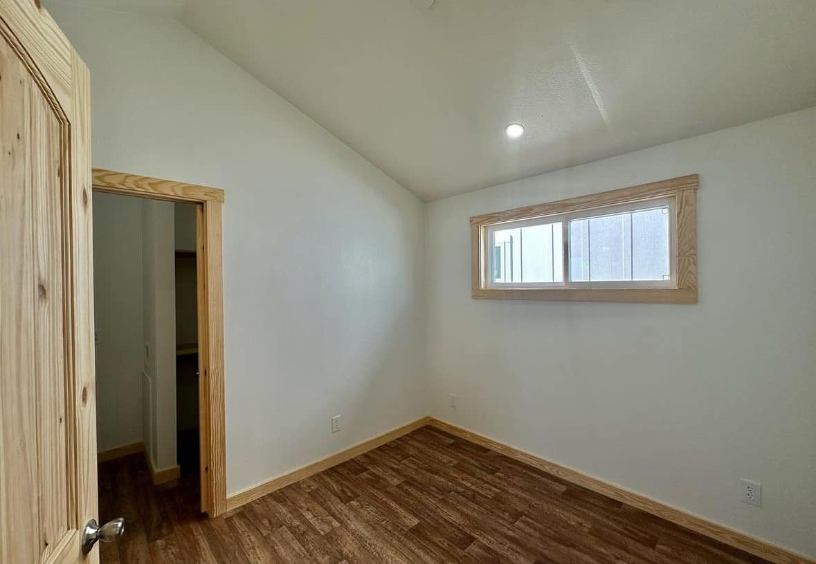 Small, empty room with white walls, wooden floor, and a high ceiling. It features a small rectangular window and an open closet. Bright, minimalistic feel.