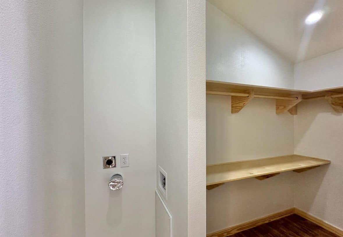 Empty white utility room corner with wooden shelves on the right, outlet and hose connections on the back wall, on a wooden floor. Modern and minimalistic.