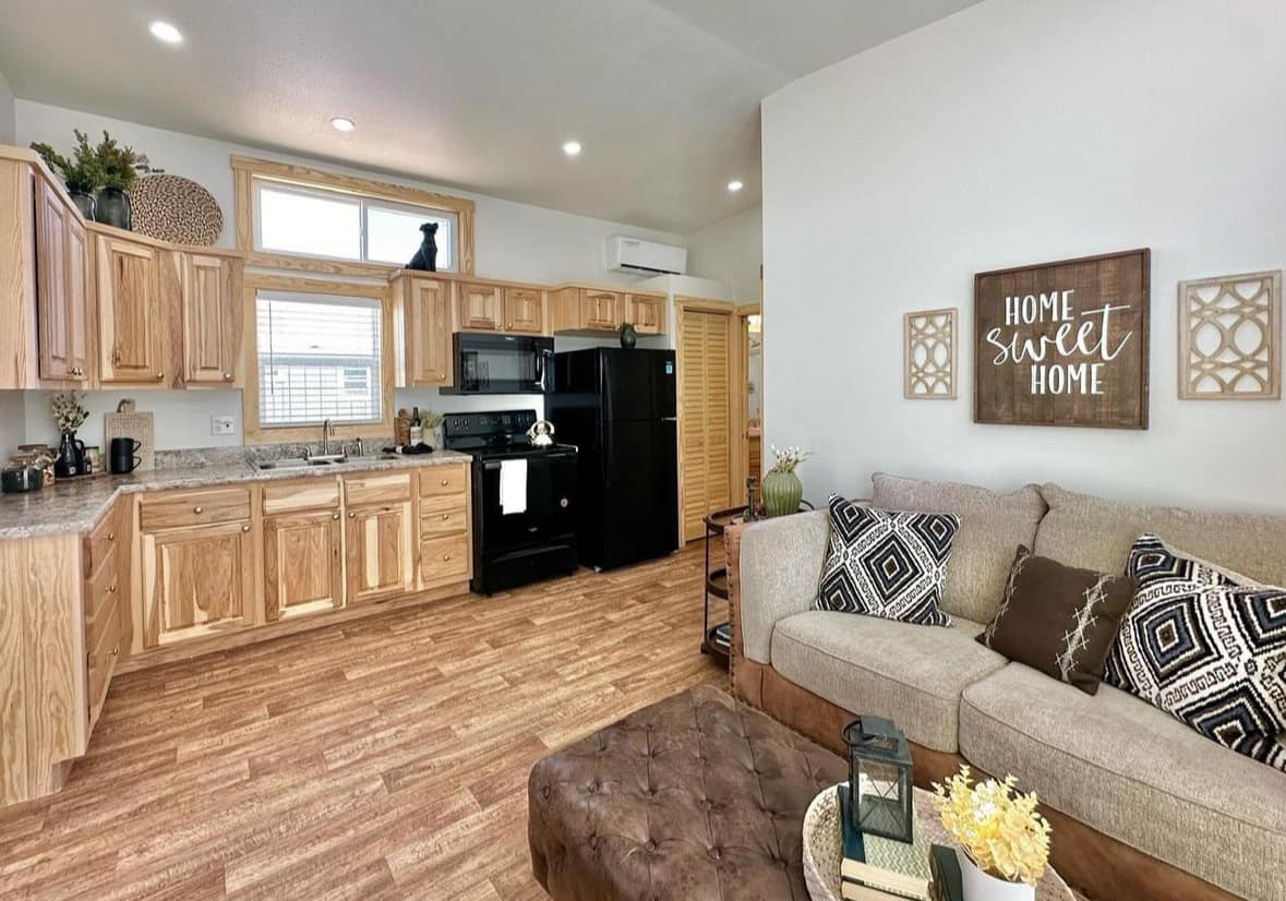 Cozy living area with a tan sectional sofa adorned with black and white pillows. Adjacent wooden kitchen features black appliances. A sign reads "Home Sweet Home," enhancing the welcoming atmosphere.