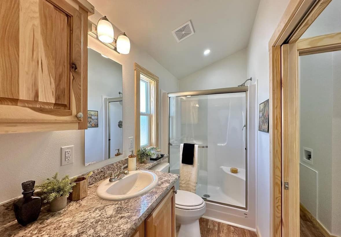 Cozy bathroom with wood cabinets, a granite counter, and a sink. A shower with glass doors is in the background. Warm lighting and potted plants add a welcoming touch.