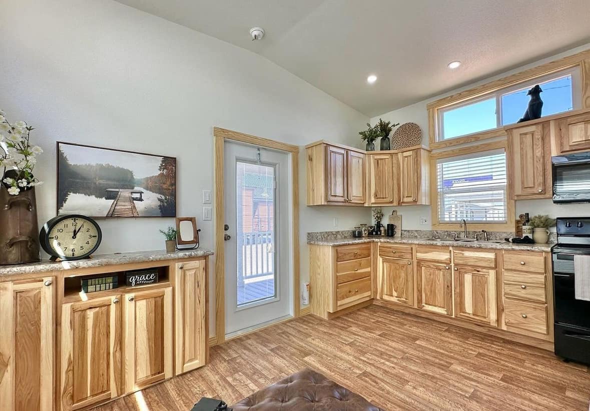 Bright kitchen with wooden cabinetry and a stone countertop. A glass door leads outside. A clock and nature art add warmth. Hardwood floors enhance the cozy feel.