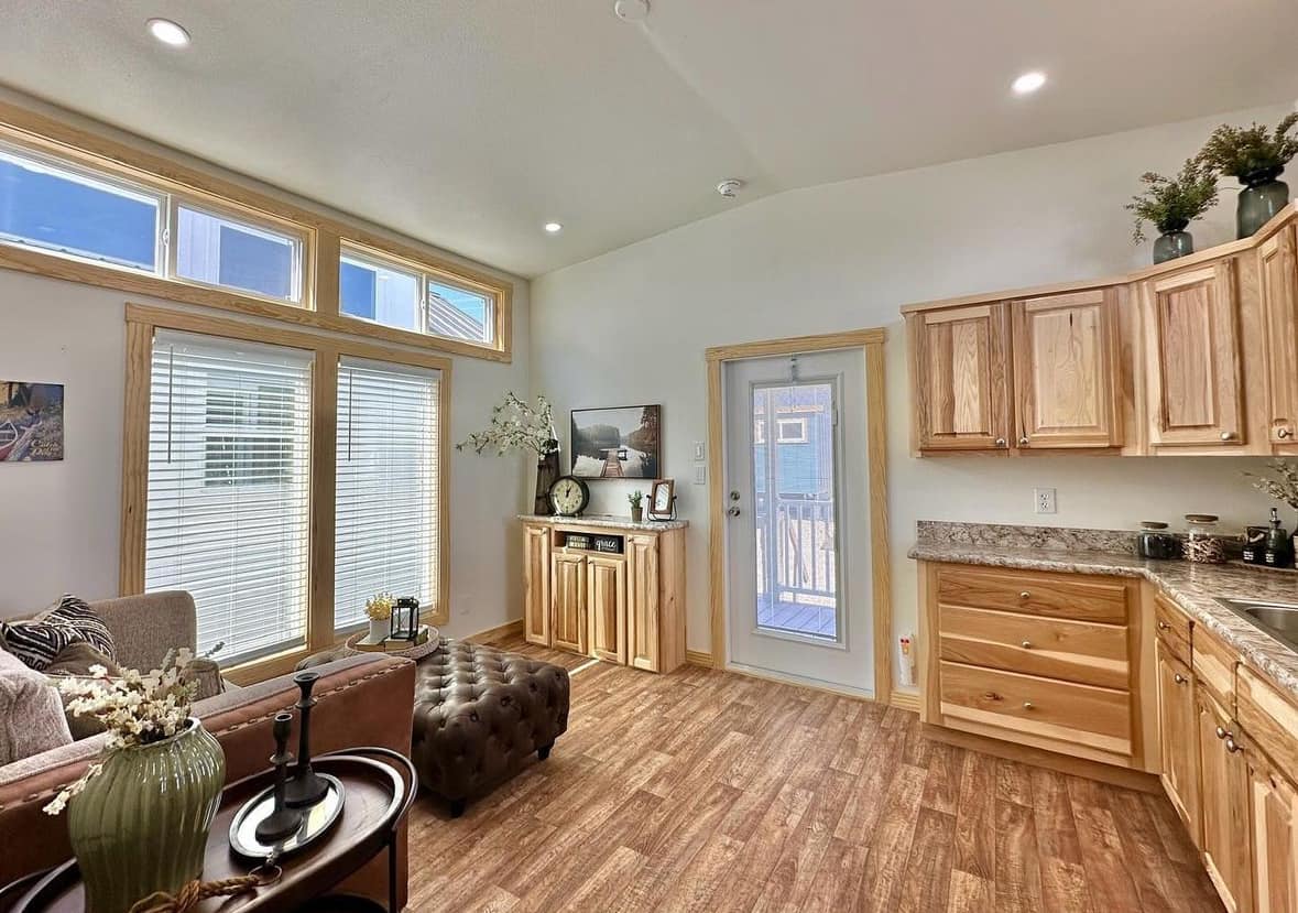 Bright, cozy living room with wooden cabinets, brown tufted ottoman, and beige sofa. Large windows and a glass door let in natural light.